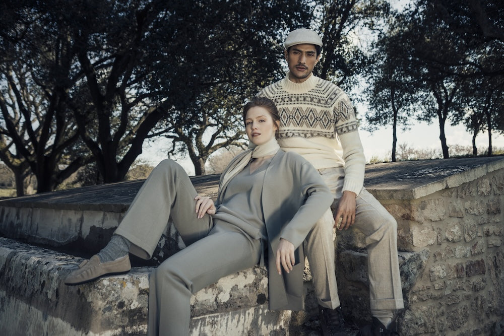 Frau in einem grauen Outfit und Mann mit einem hellen Musterpullover, draußen auf einer Steinmauer sitzend.