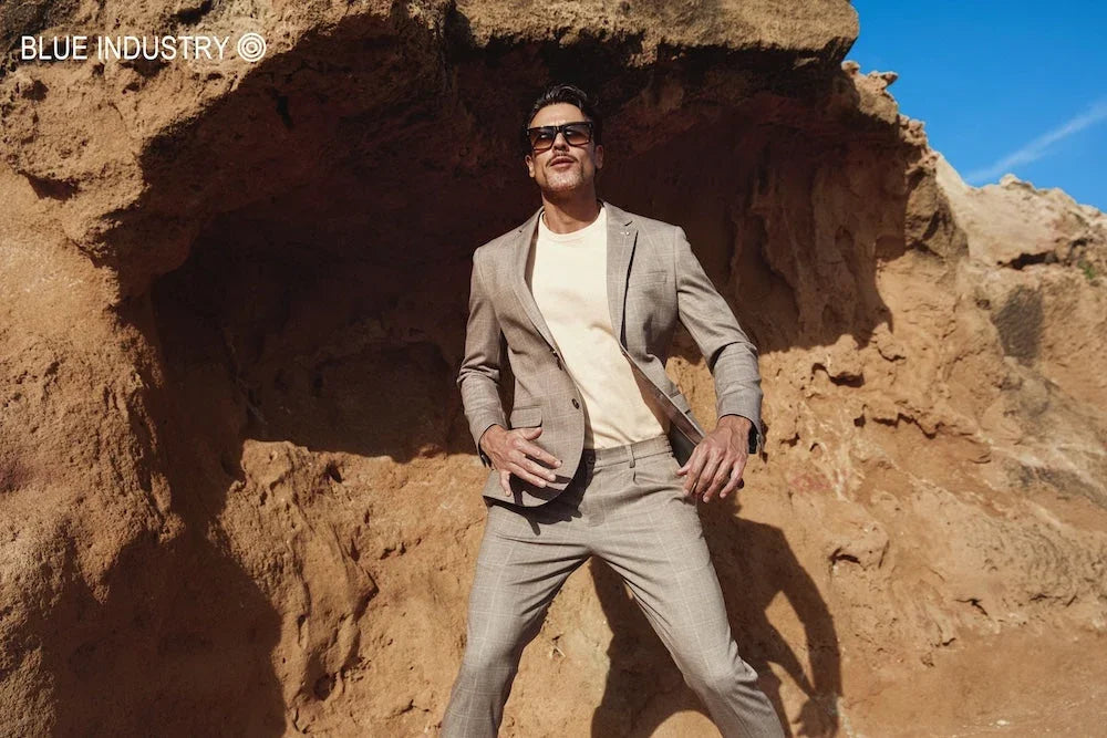 Man in een beige geruit herenkostuum met beige t-shirt en zonnebril voor een rotswand.